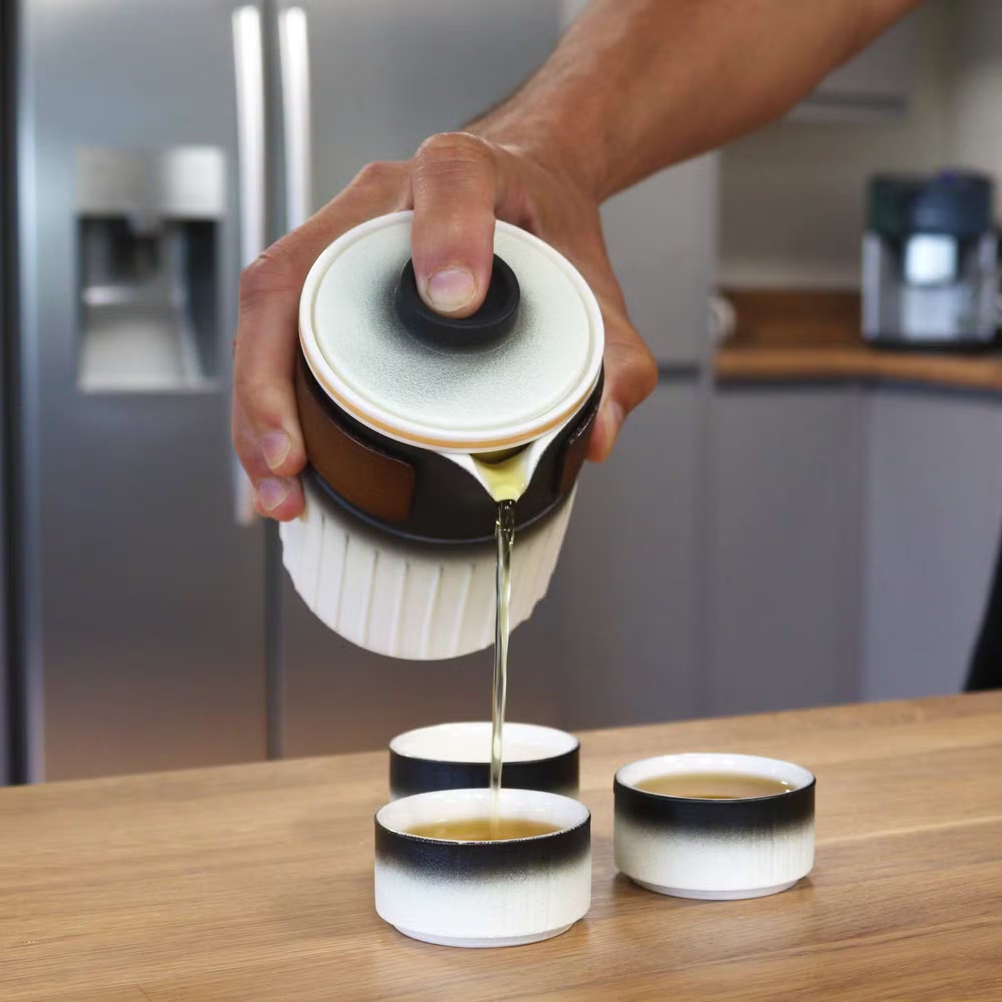 Person pouring liquid from a white container with black accents into three small cups on a wooden surface.